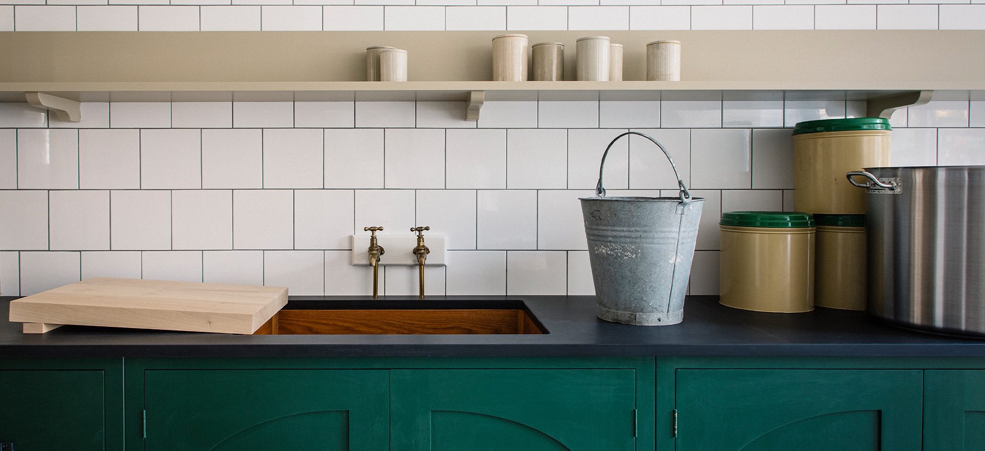 A bespoke kitchen with green cabinetry, white tiled backsplash, brass taps, and a metal bucket on a dark countertop.