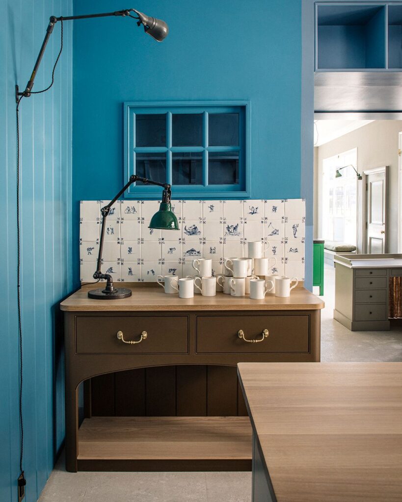 A kitchen with blue walls features a brown wooden cabinet, a green desk lamp, white mugs, and a white tile backsplash with blue patterns.