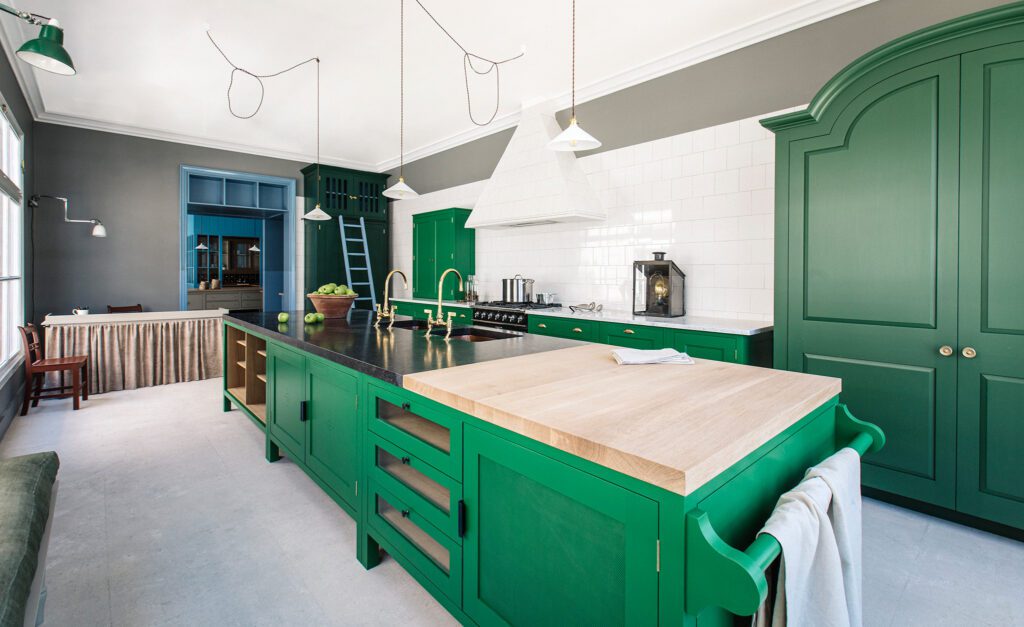 A spacious kitchen features vibrant green cabinetry, a large wooden island, white tiled backsplash, and gray walls, with pendant lights and a blue accent wall in the background.