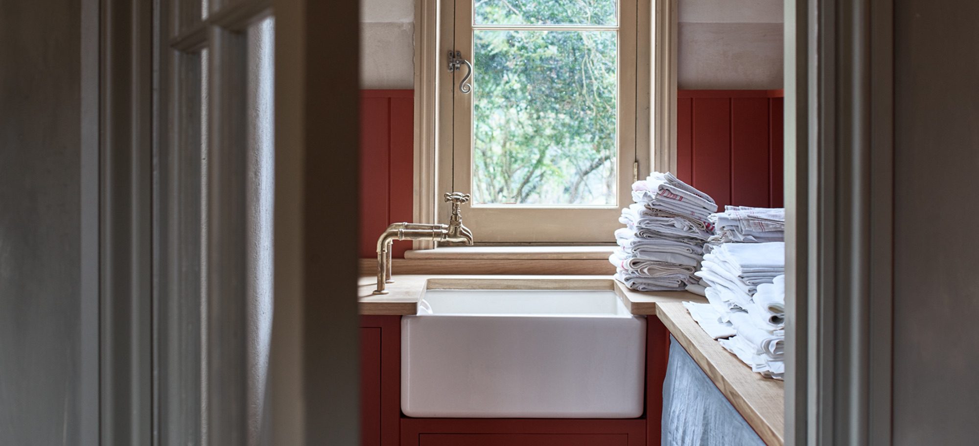 A kitchen with a farmhouse sink, brass faucet, red cabinetry, and a stack of linens by a window overlooking greenery.