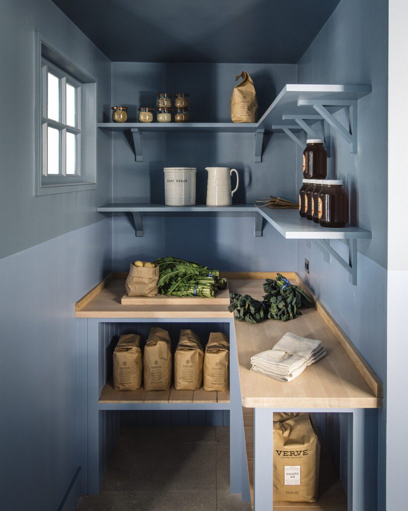 A pantry with blue walls and shelves, featuring neatly arranged jars, bags, and fresh greens on a wooden countertop.