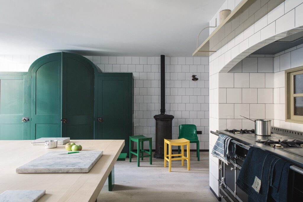 A kitchen with green cabinetry, white tiled walls, a black stove, and colorful chairs, featuring a light wood table with marble accents.
