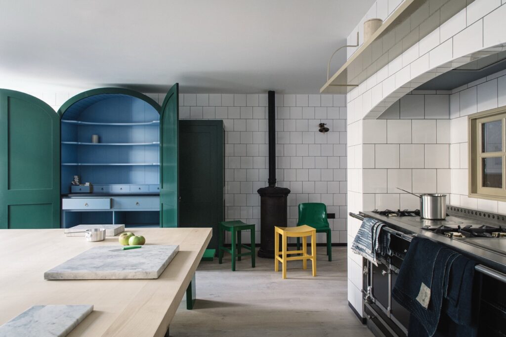 A kitchen with white tiled walls features a teal arched cabinet, a black stove, green and yellow chairs, and a large wooden island with marble accents.