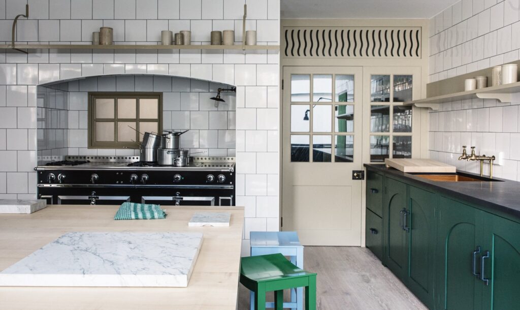 A kitchen with white tiled walls, a black stove, green cabinetry, a light wood island with a marble top, and a cream door with glass panels.