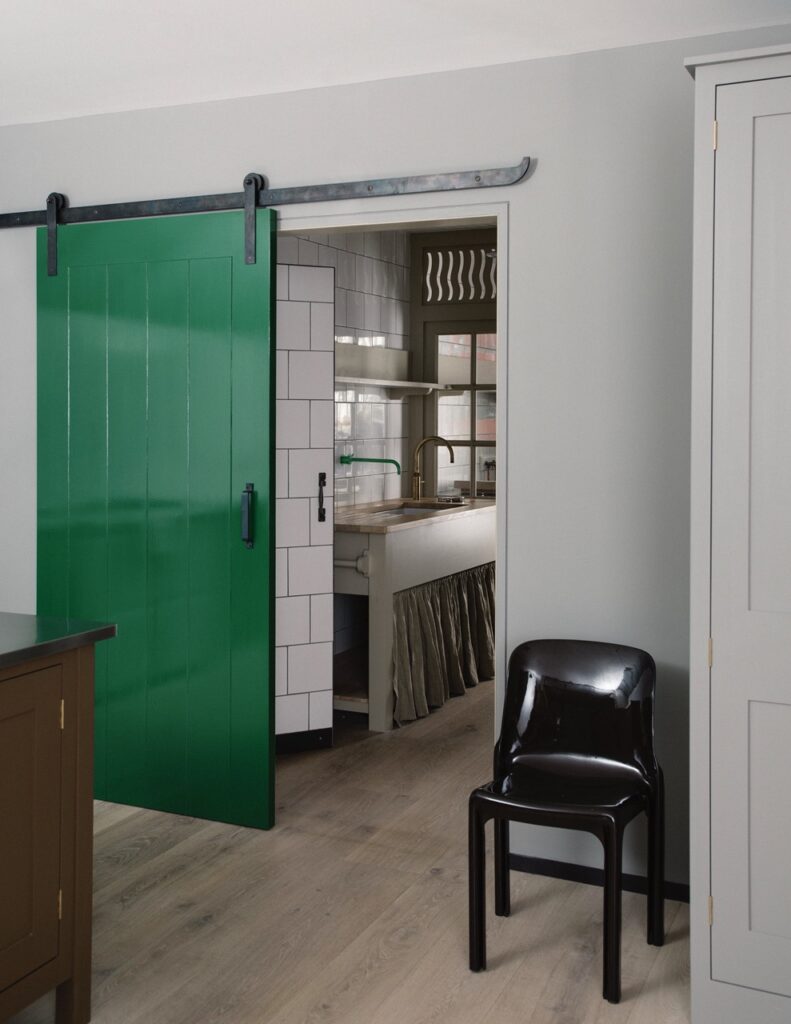 A room with a green sliding door opens to a kitchen featuring white tiled walls, a farmhouse sink, and a black chair against light gray walls and wooden flooring.
