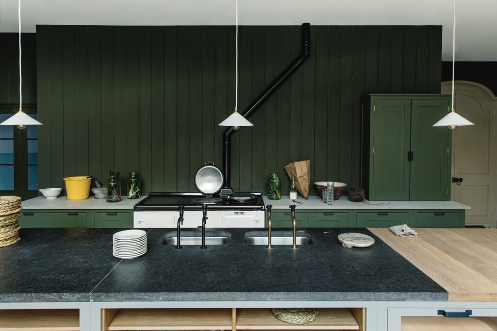 A kitchen with dark green cabinetry, a black countertop, and wooden accents features pendant lights and a classic stove, blending traditional and modern elements.