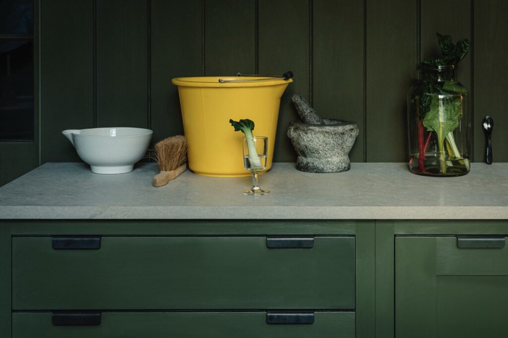 A kitchen countertop with dark green cabinetry features a white bowl, a yellow bucket, a brush, a glass with celery, a stone mortar and pestle, and a jar with vegetables against a dark green paneled wall.