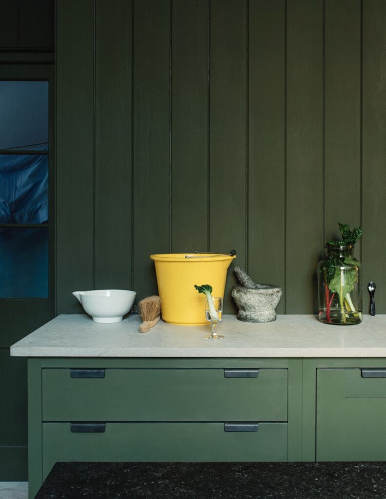 A kitchen with dark green cabinetry features a light countertop adorned with a yellow bucket, white bowl, mortar and pestle, and a jar of vegetables.