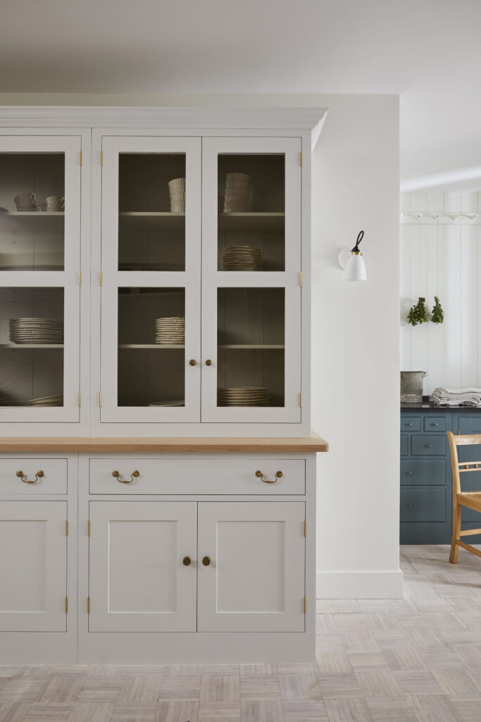 A white kitchen hutch storing plates and cups, teal cupboard in back of room with wood chair