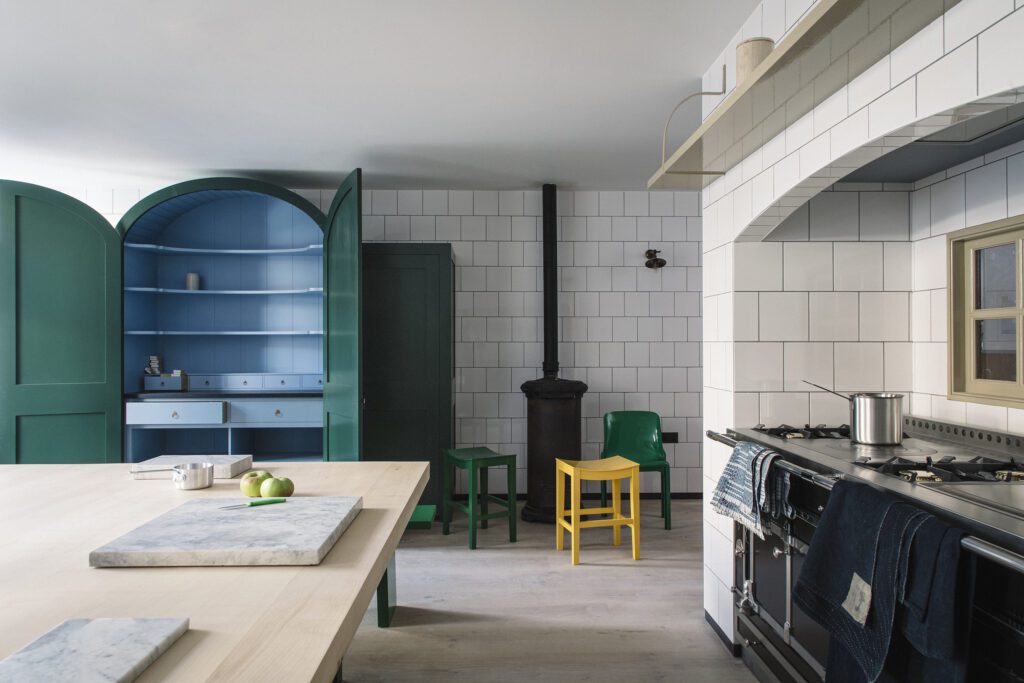 A kitchen with white tiled walls features a large stove, a light wood island, a blue and green open cabinet, green and yellow chairs, and a black stove pipe.