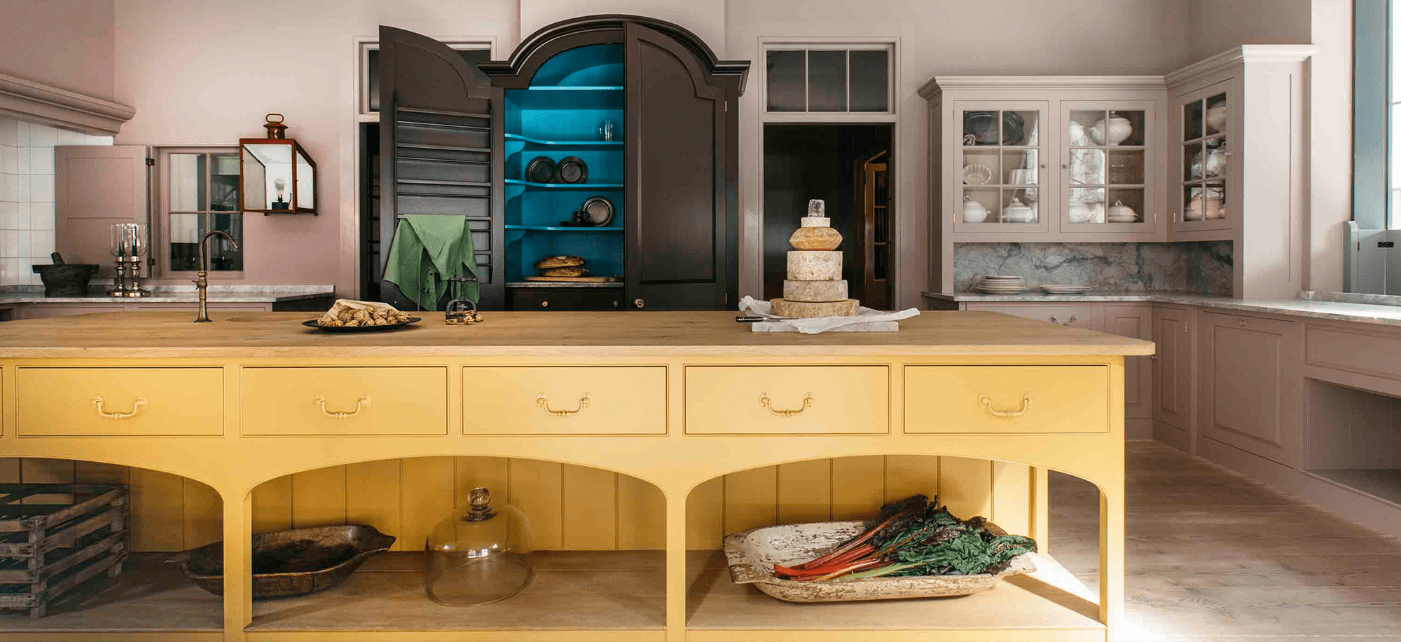 A kitchen with a yellow island, dark cabinetry with a blue interior, and a display of cheeses and vegetables.