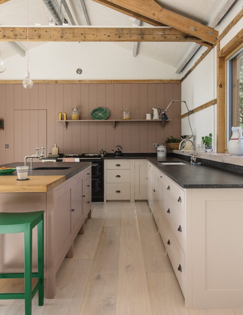 a converted barn kitchen in a mix of creamy colors, muddy lavender wall paneling, blush lower cabinets, and matching island with a wood countertop and green stool