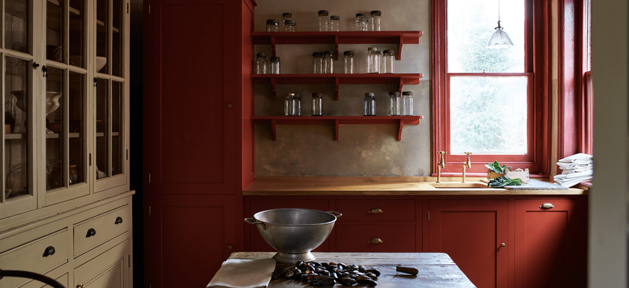 A rustic kitchen with deep red cabinetry, open shelves holding glass jars, a wooden countertop, and a metal colander on a wooden table.