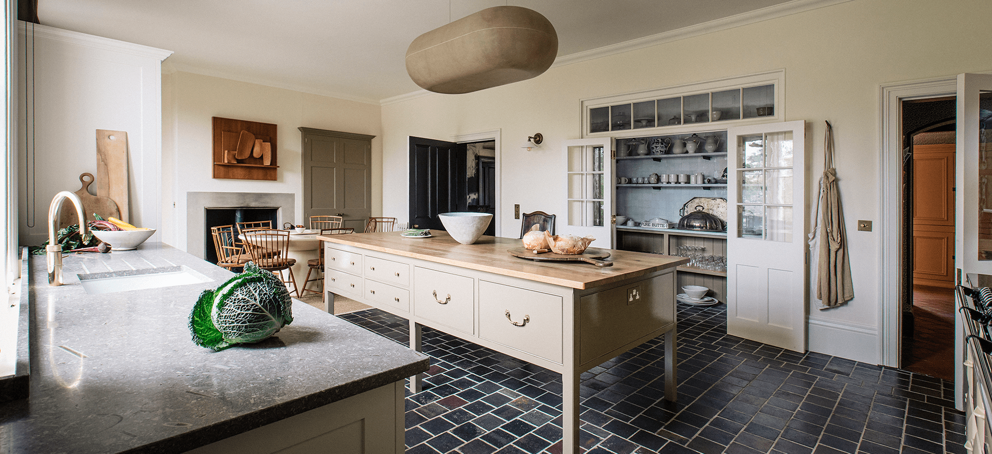 A spacious kitchen with a large wooden island, cream cabinetry, dark tiled floor, and a soft beige pendant light, featuring classic Georgian-inspired detailing.