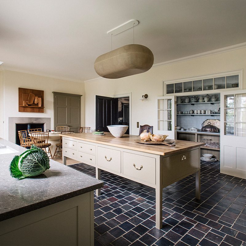 A spacious kitchen features a large beige island with wooden countertop, surrounded by dark tiled flooring, white cabinetry, and a dining area with wooden chairs, all under a unique oval light fixture.