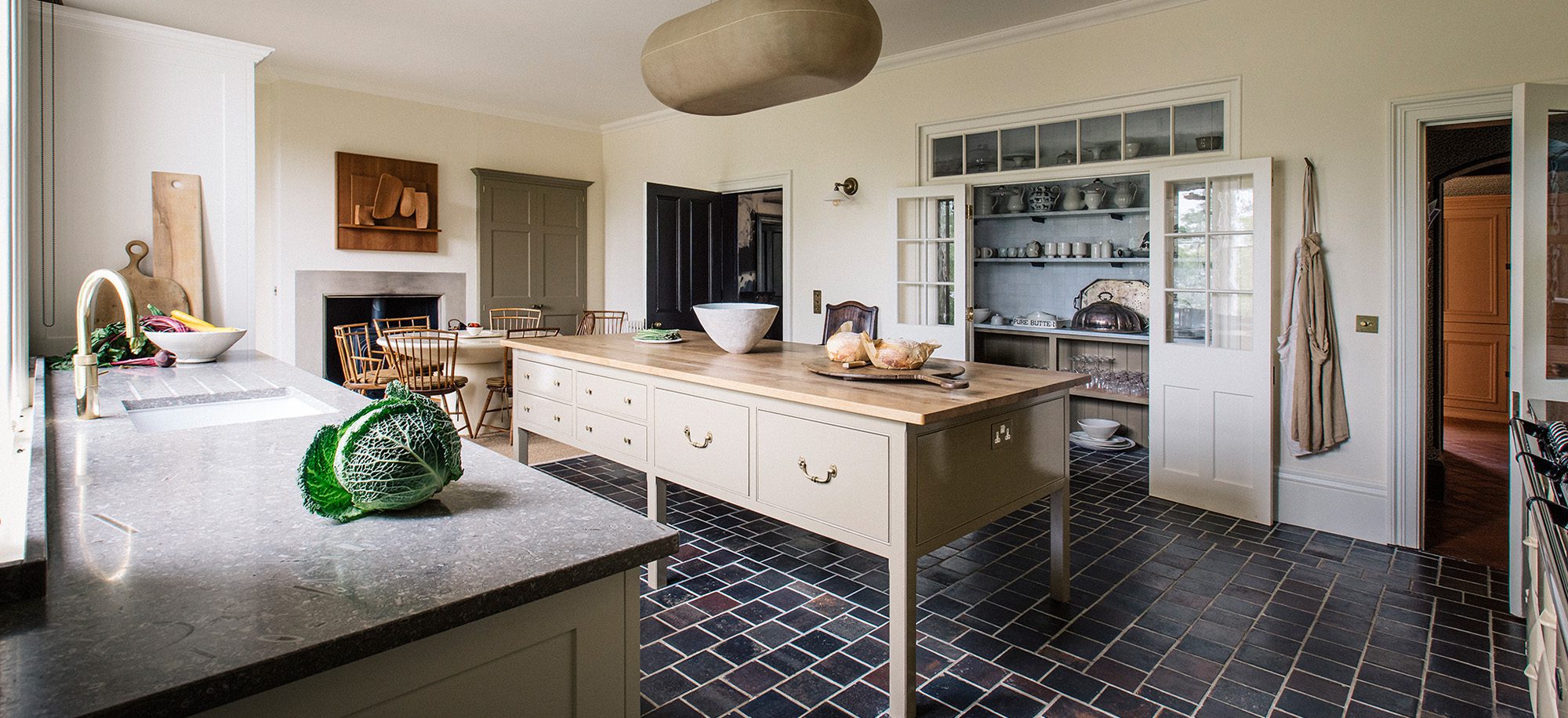  A spacious kitchen with cream cabinetry, a wooden island, dark tiled floor, and a large windowed pantry, featuring classic Georgian-inspired design elements.
