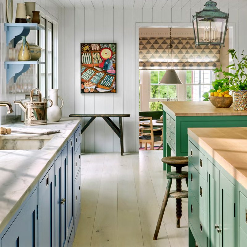 A bright kitchen with blue and green cabinetry and marble countertops, leading to a dining area with patterned curtains and a large window.