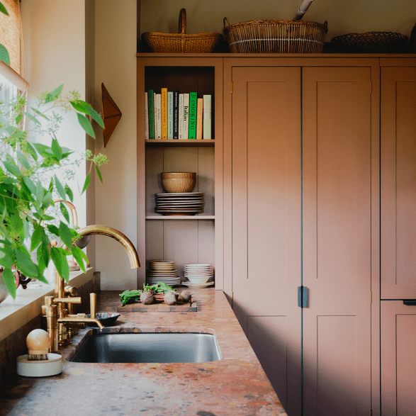 A warm kitchen features bespoke cabinetry in muted pink, a marble countertop, brass fixtures, and open shelving with neatly arranged dishes and books.